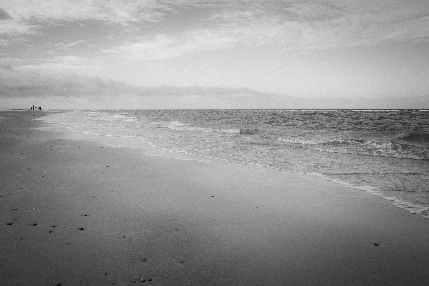 Black and white serene beach scene with gentle ocean waves and two distant figures on the shoreline.