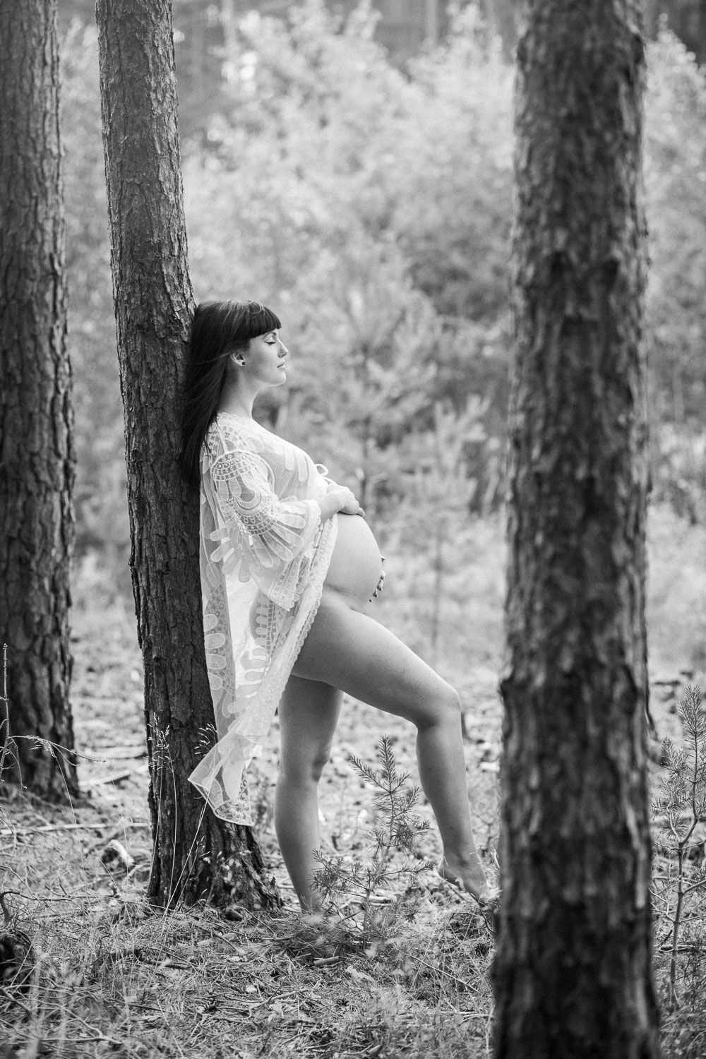 Black and white artistic portrait of a pregnant woman in lace robe leaning against a tree in a forest.