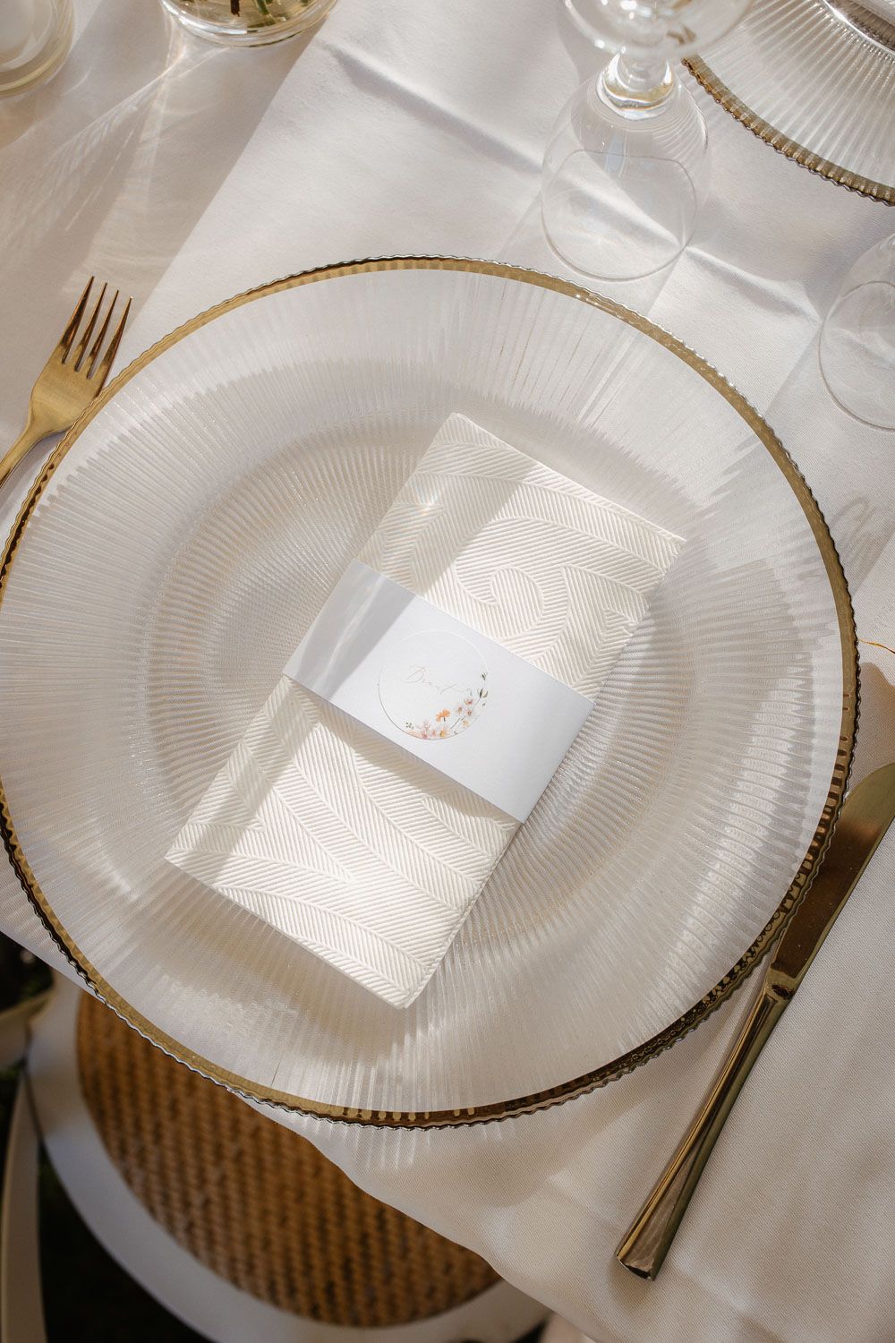 Elegant wedding table setting with a glass charger plate, gold cutlery, and a folded napkin with a floral card.