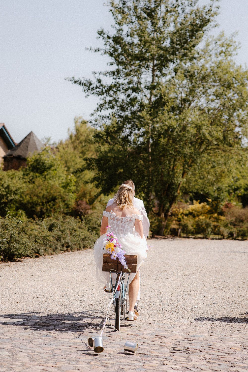 Bride in short white tulle dress riding a decorated bicycle on a sunny gravel path outdoors.