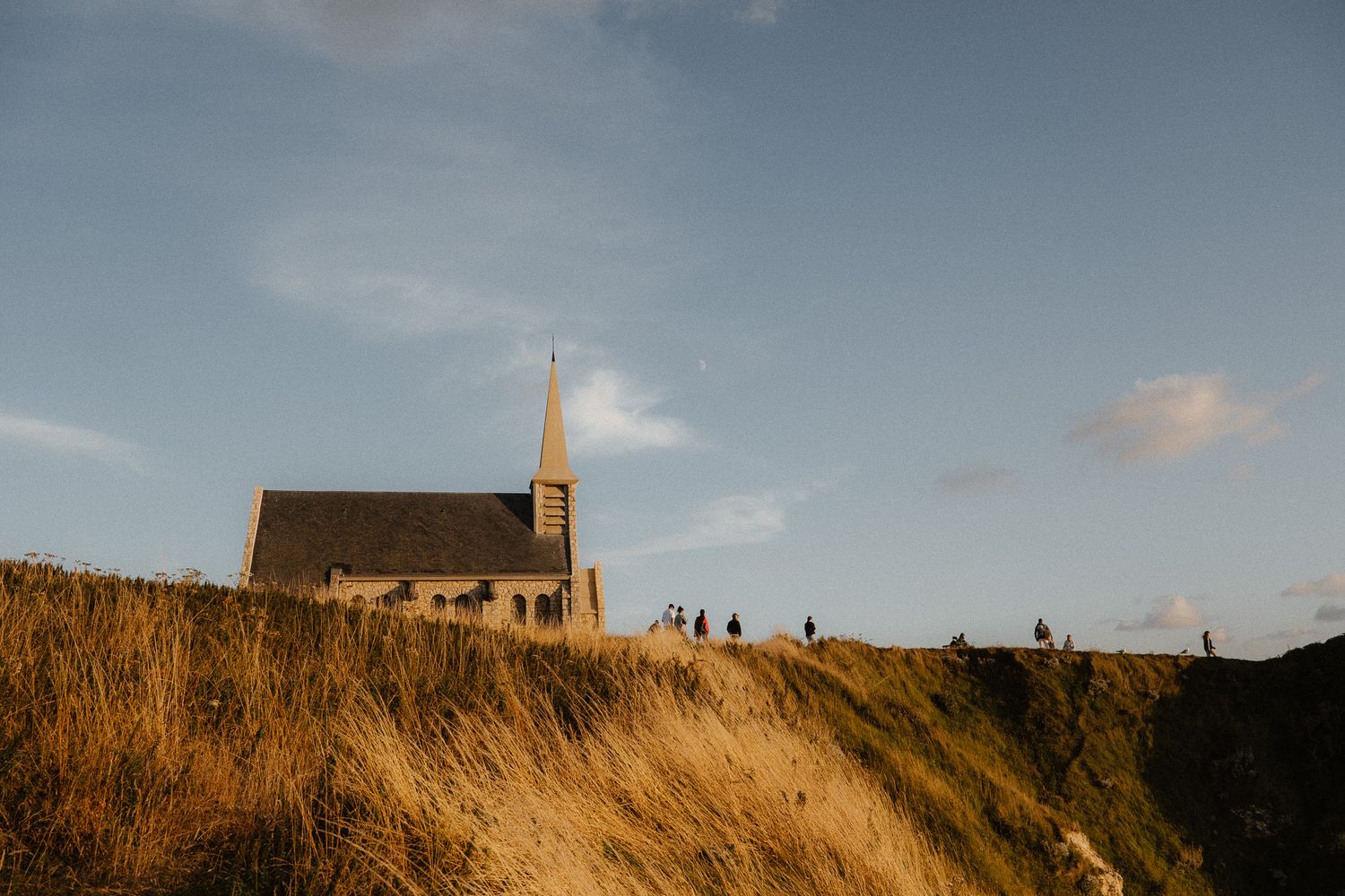 Historic stone church with steeple on a golden grassy cliff edge under a blue sky in Normandy, France.