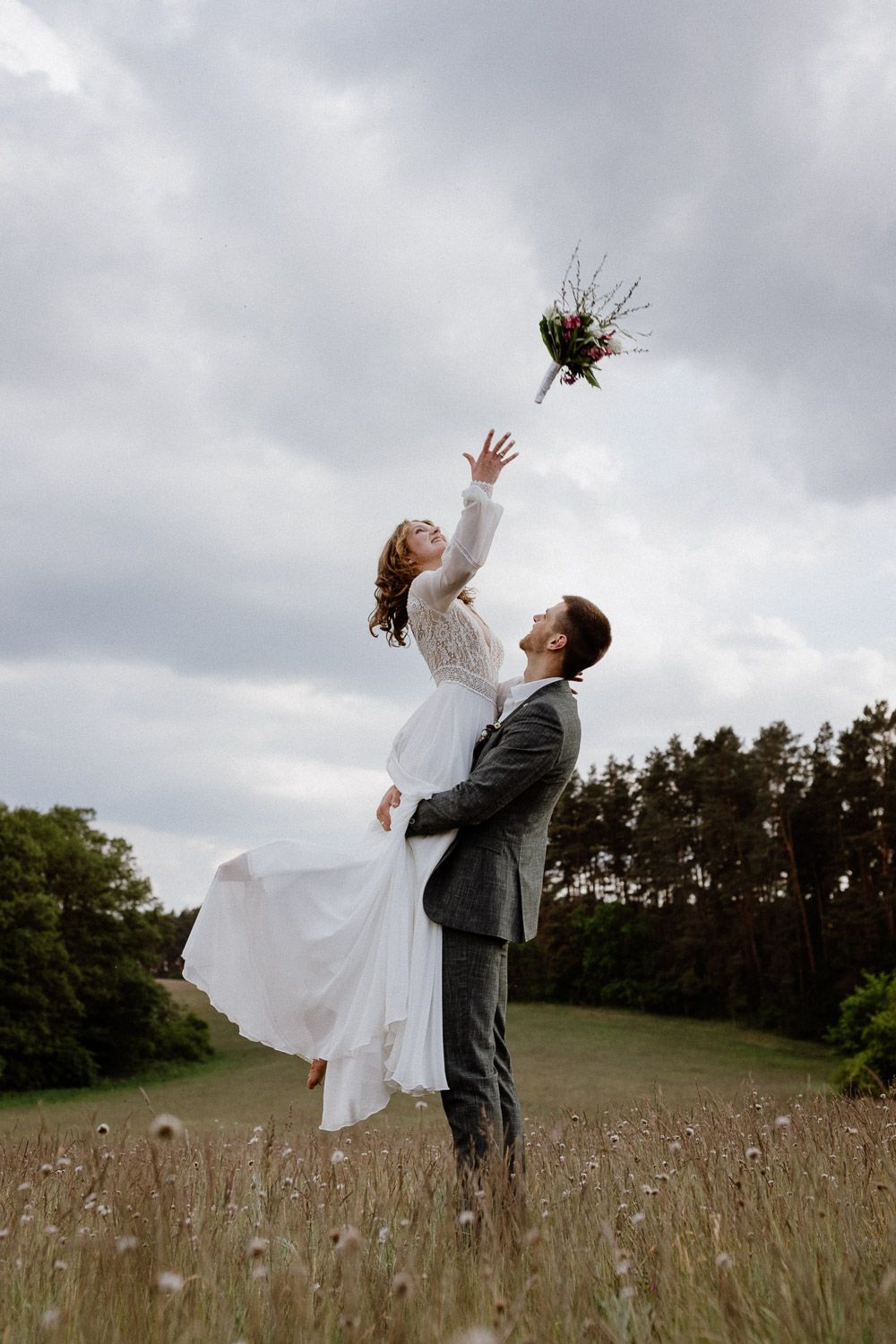 Groom lifting bride in white dress as she tosses bouquet into cloudy sky in an open field.