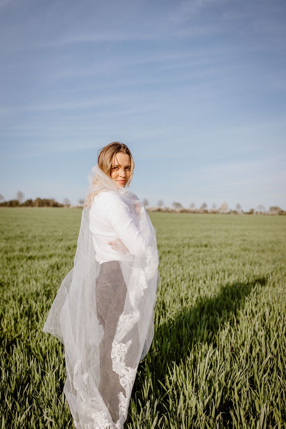 Bride in white dress and lace veil standing in a green field under a blue sky.