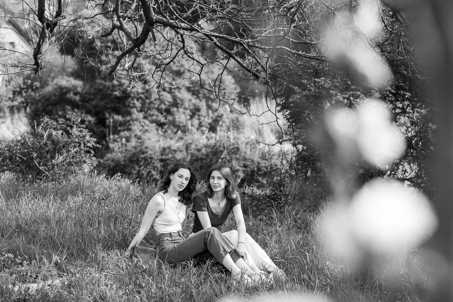 Two women sitting together in a grassy field under tree branches in a black and white outdoor portrait.