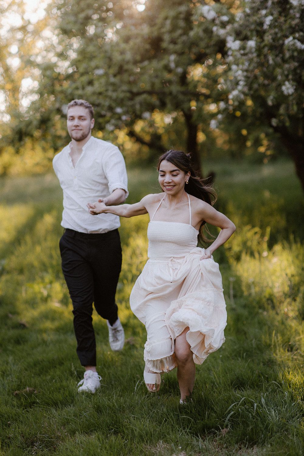 A joyful couple runs hand in hand through a sunlit orchard, bride in a flowing white dress.
