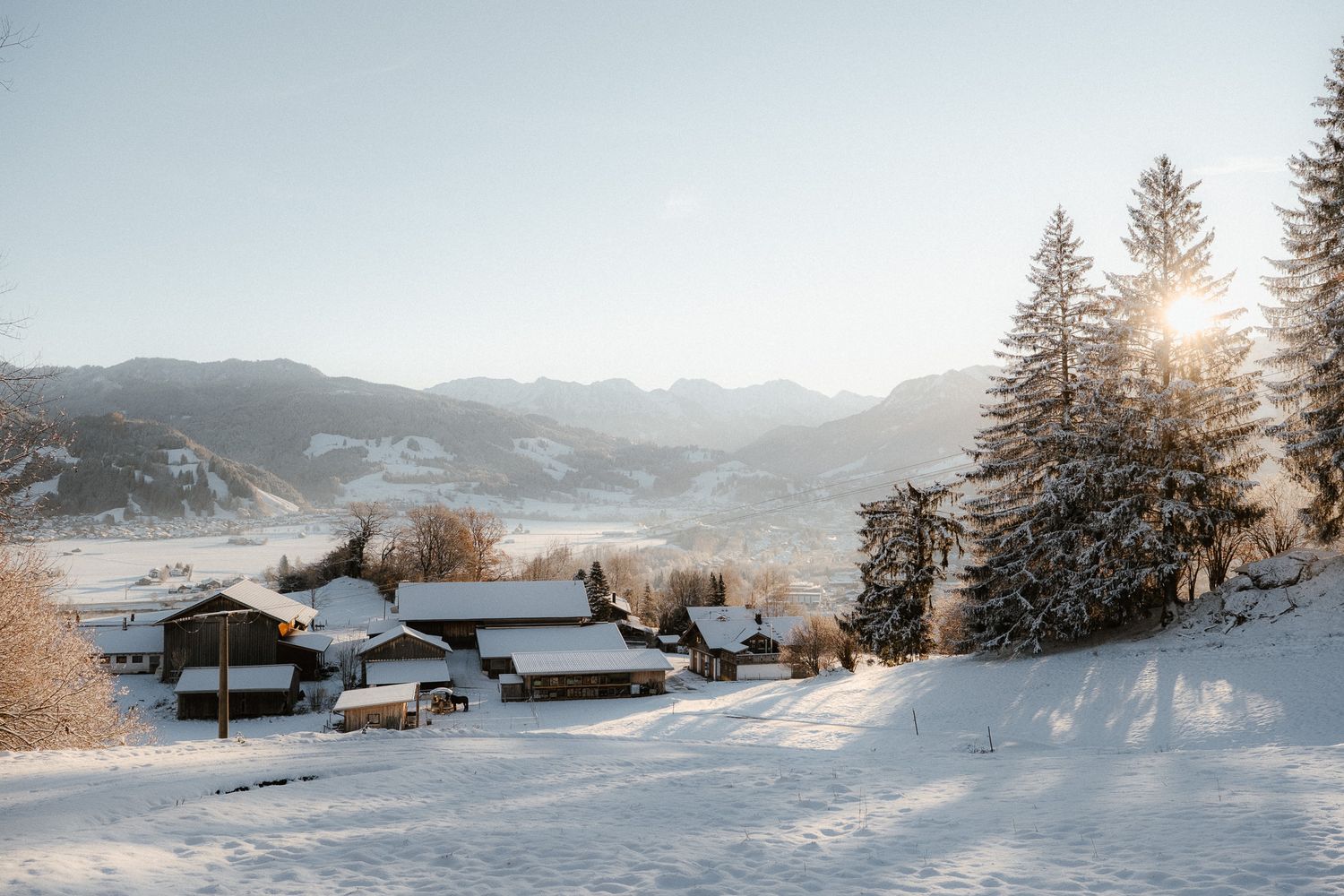 Snowy Alpine village at sunset with snow-covered chalets, pine trees, and mountain valley backdrop.