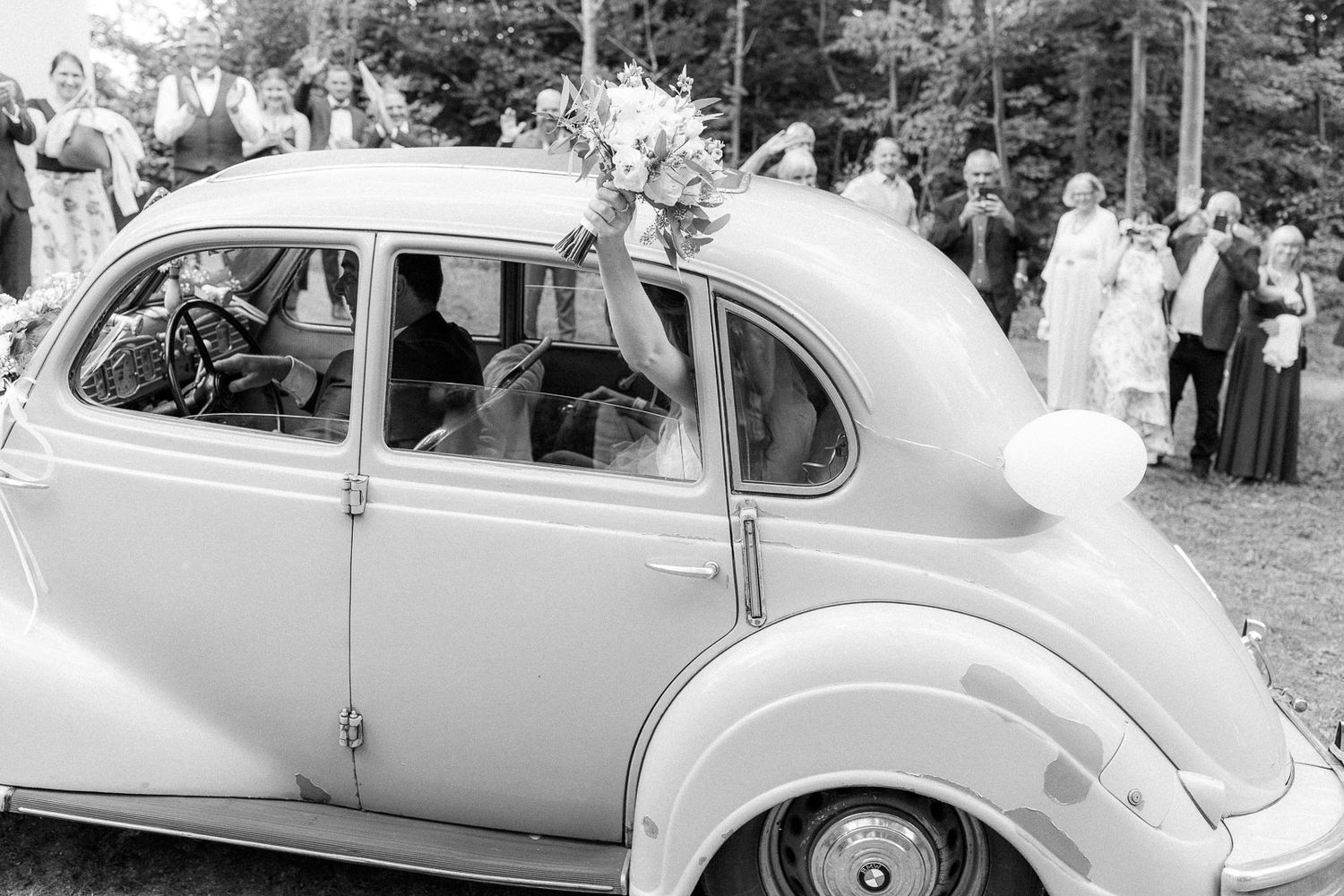 Black and white image of a bride holding a bouquet inside a vintage Volkswagen Beetle at a wedding.