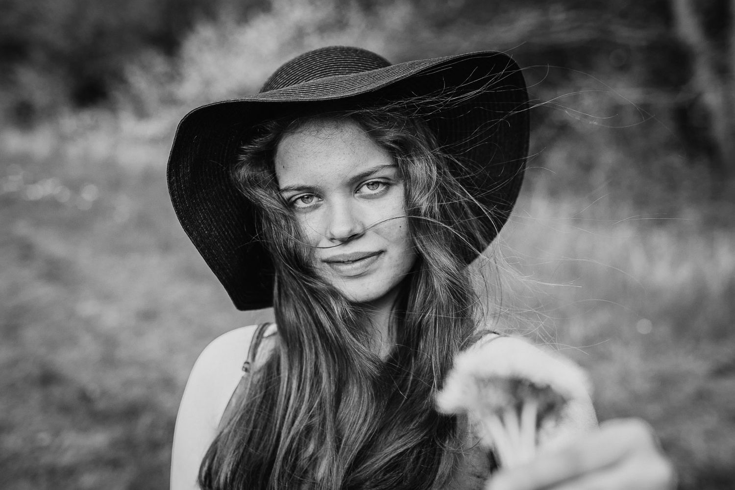 Black and white portrait of a young woman wearing a wide-brim hat in a natural outdoor setting.