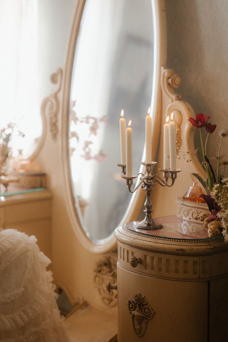 Vintage vanity with ornate oval mirror, silver candelabra with lit candles, and floral decor.