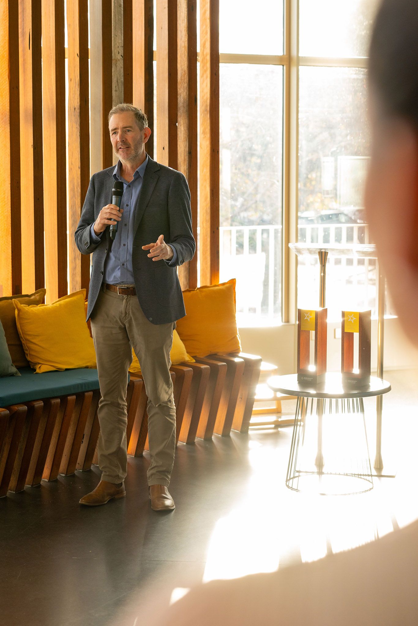 Man in blazer speaking into microphone at modern venue with yellow cushions and wooden slat walls, event photography