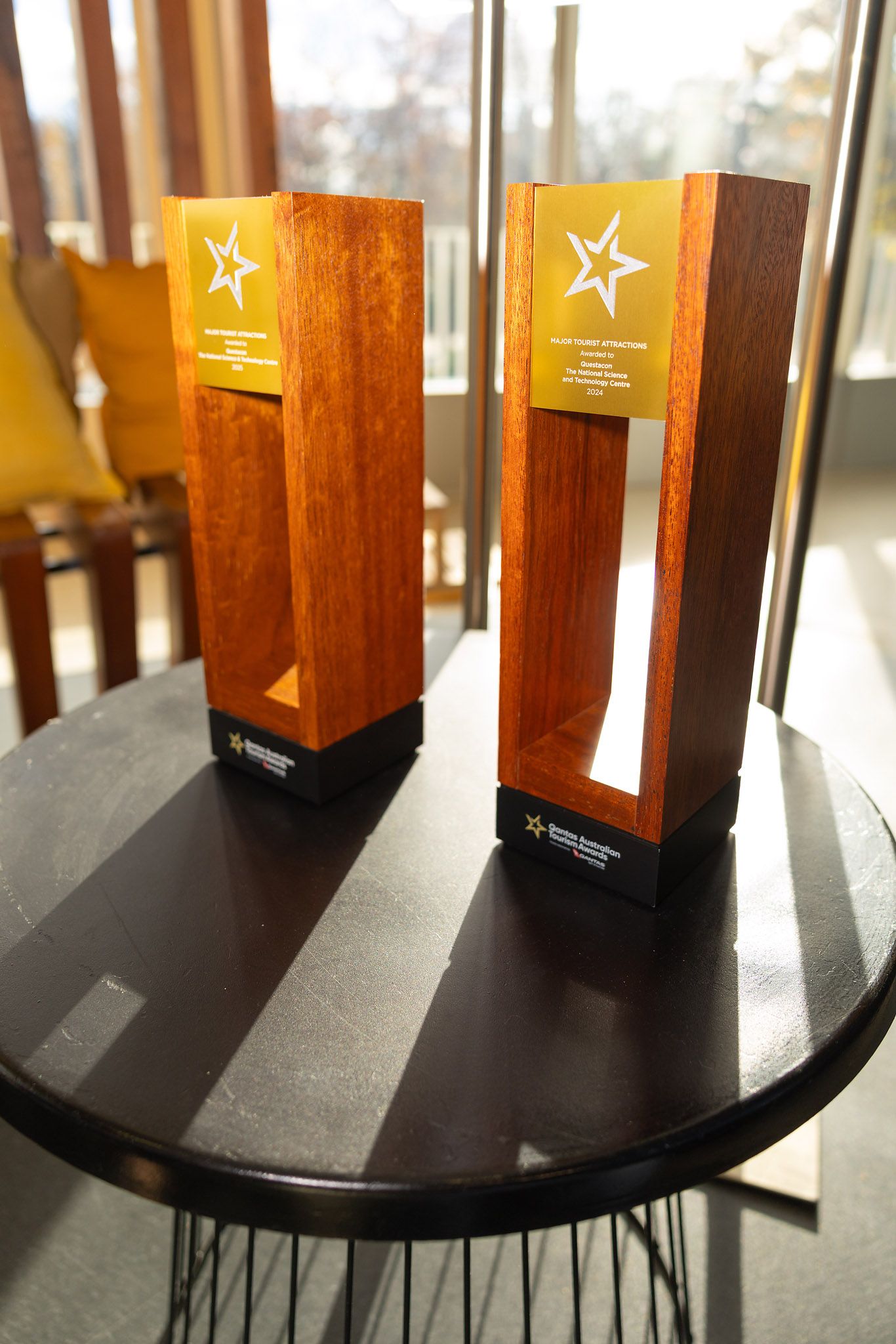 Two wooden trophy awards with gold star emblems displayed on a dark round table near a sunlit window, Canberra Tourism Industry  Award
