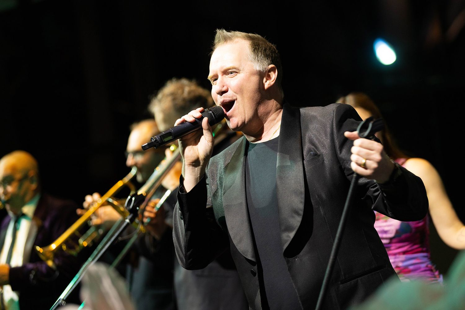 Male singer performing passionately on stage with a band, wearing a black suit and holding a microphone.