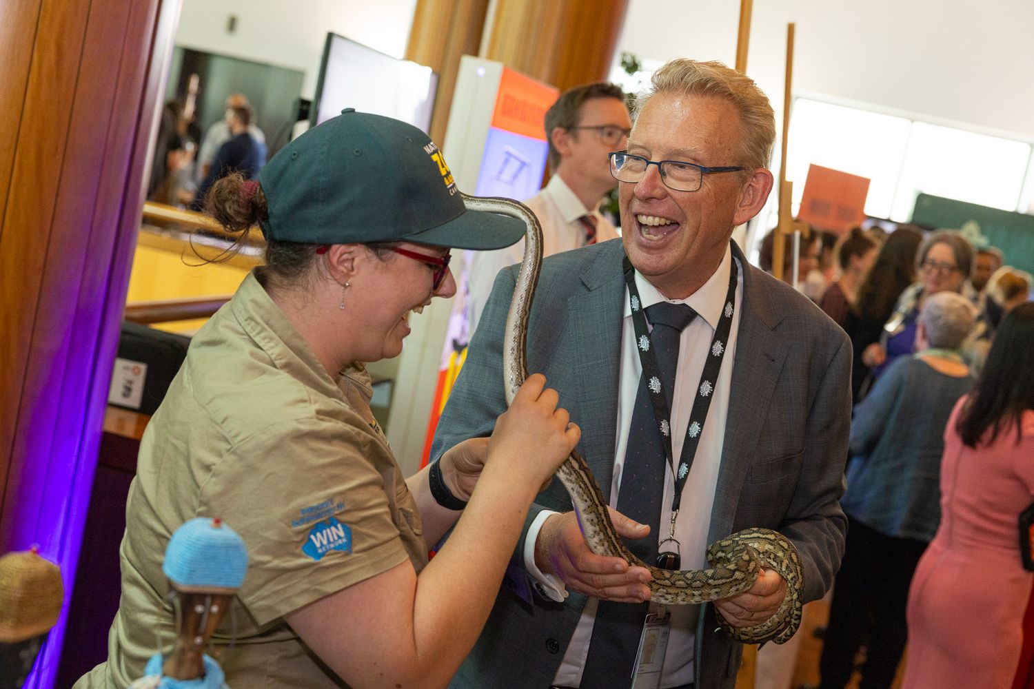 Zookeeper and man in suit laughing while handling a snake at an indoor event at Parliament House, Canberra.