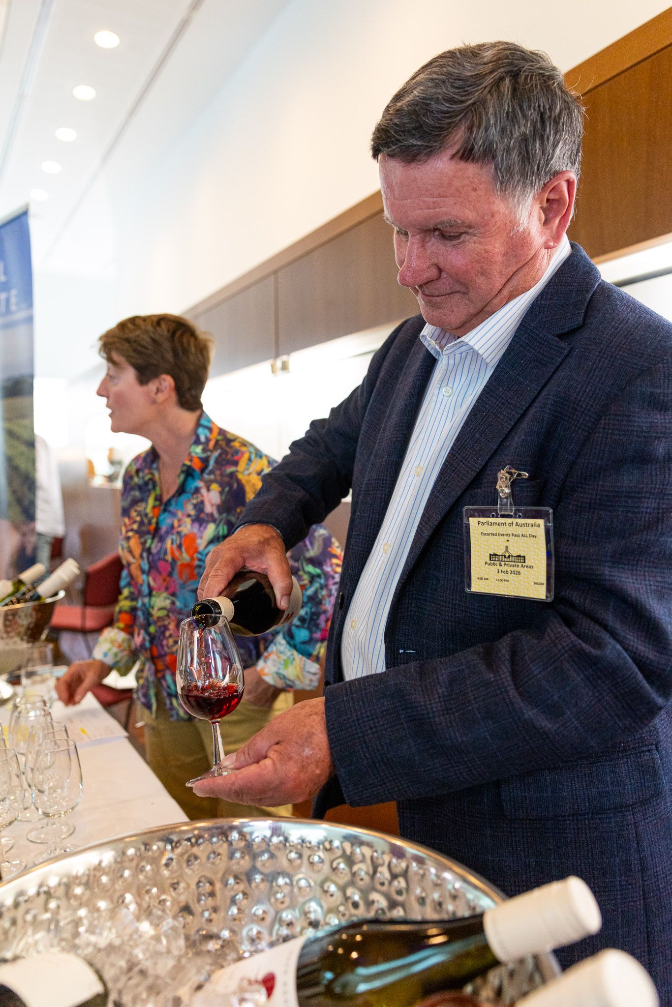 Man in blazer pouring red wine at an elegant tasting event, with guests and wine bottles visible in background.