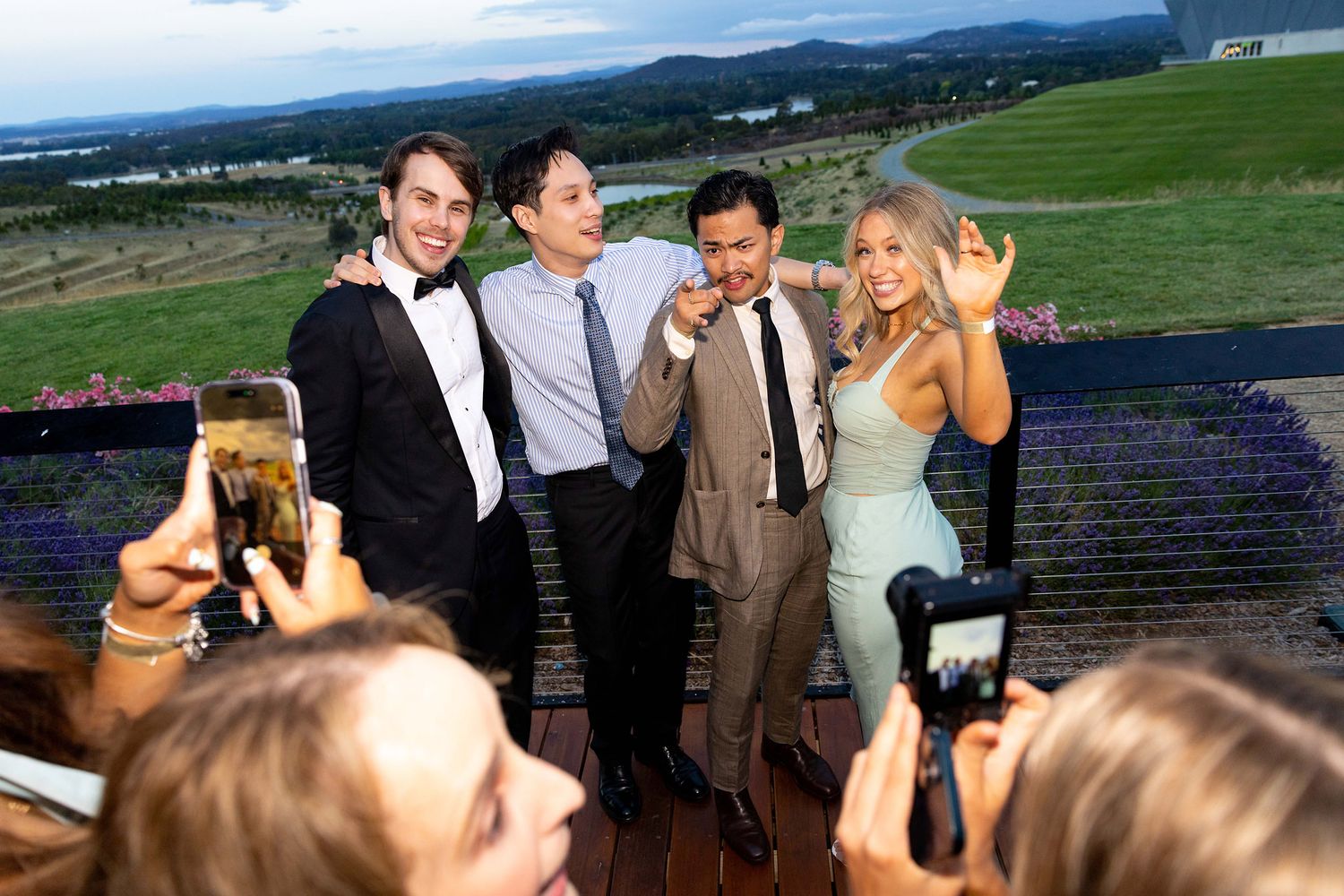 Four elegantly dressed guests pose for photos at an outdoor event with scenic green hills in the background.