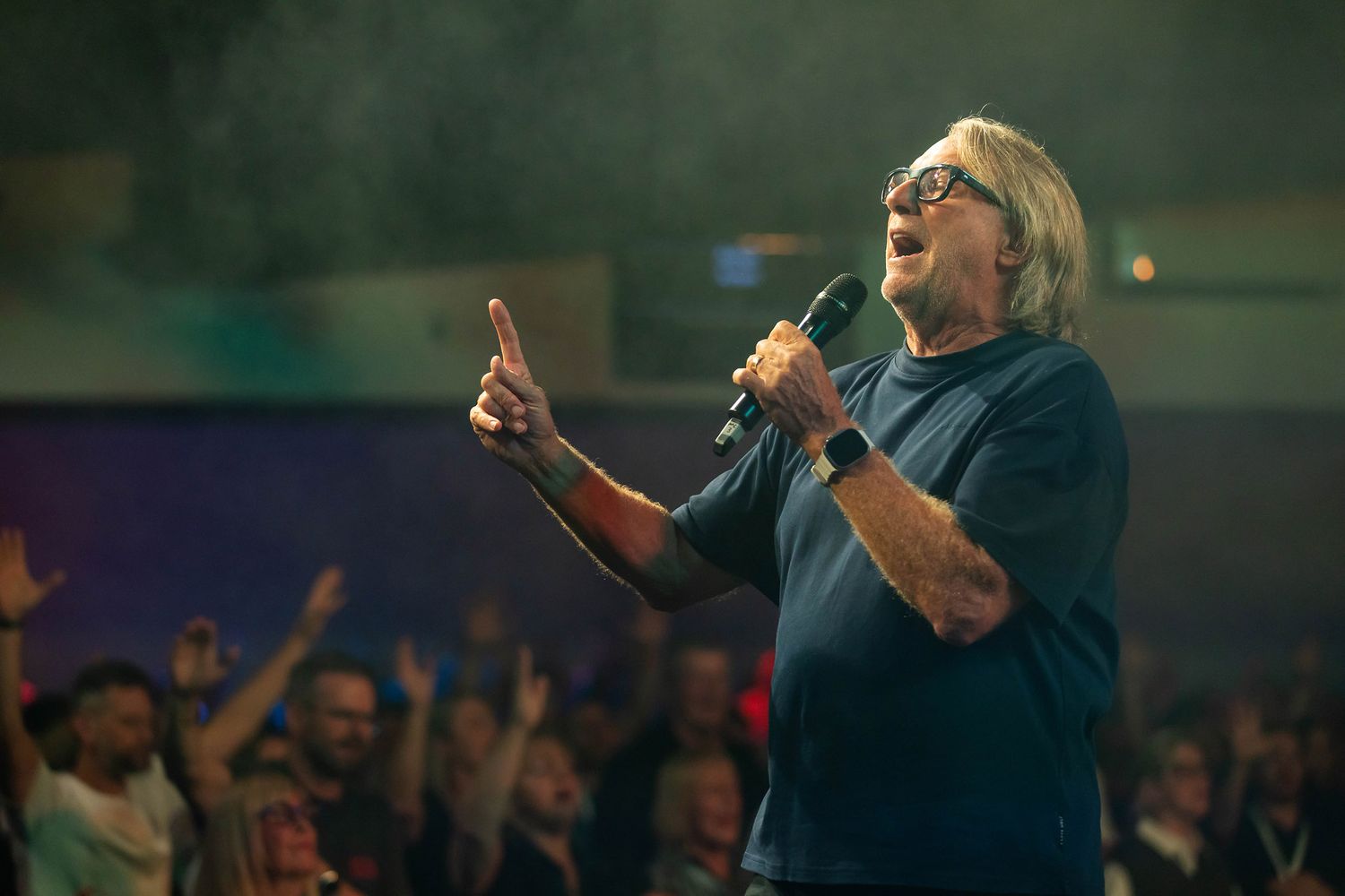 Man with long white hair speaks into microphone on stage, pointing upward, with cheering crowd behind him.