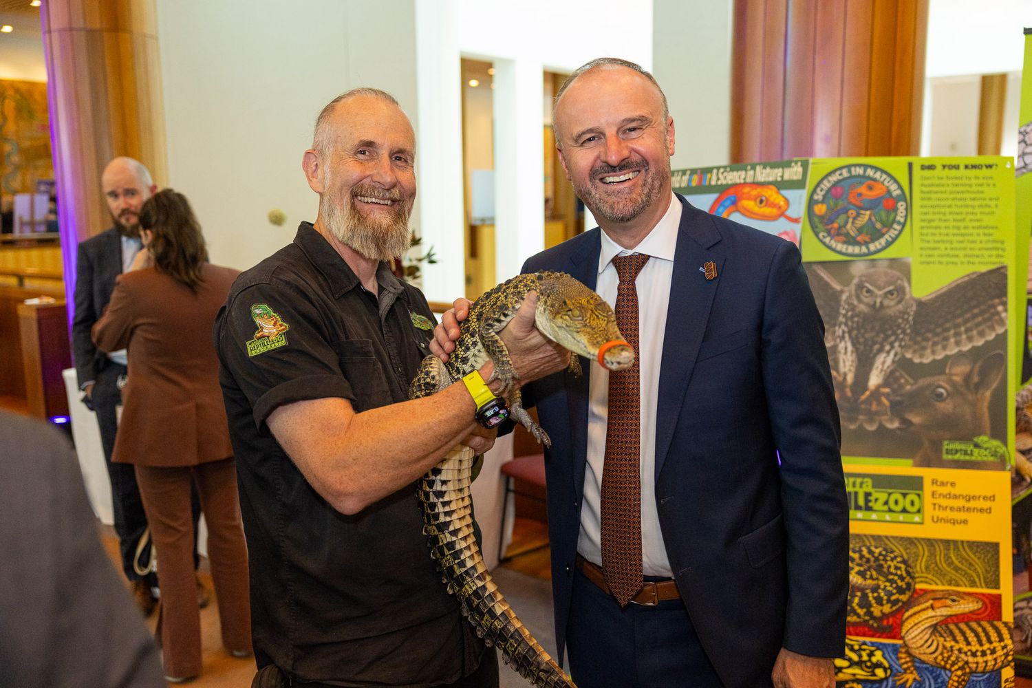 A zookeeper and a politician smile holding a large lizard at an indoor wildlife education event with colorful reptile display boards.