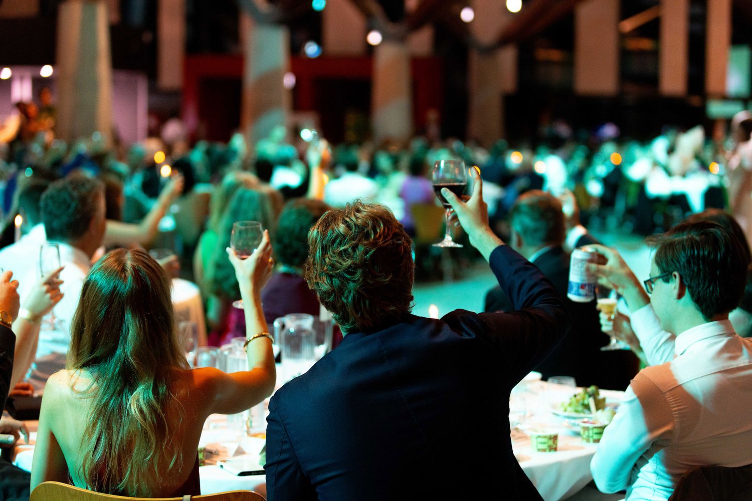 Crowd of guests raising glasses in a toast at a formal gala dinner event with colourful ambient lighting.