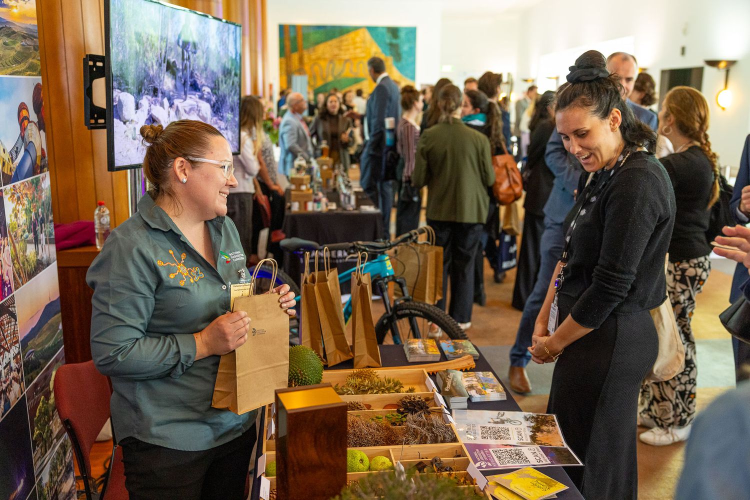 Two women conversing at an indoor expo booth displaying nature photos and educational materials at a busy tourism event, Parliament House