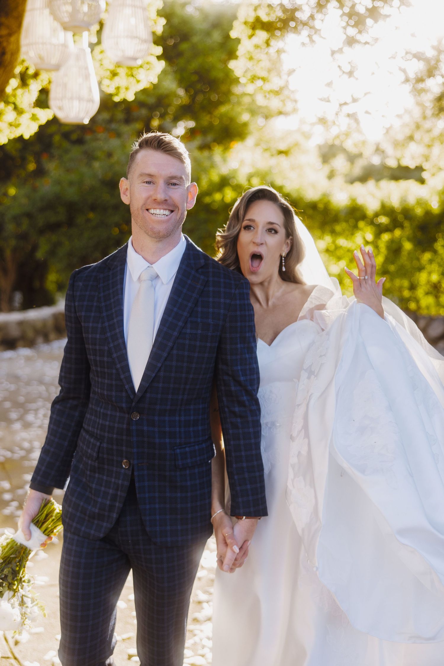 A joyful wedding couple poses together outdoors in sunlight with the bride making a playful expression.