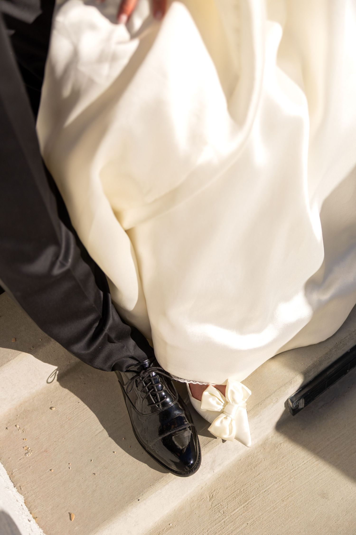 A close-up shot of a shiny black formal shoe next to flowing white wedding dress fabric on a light floor.