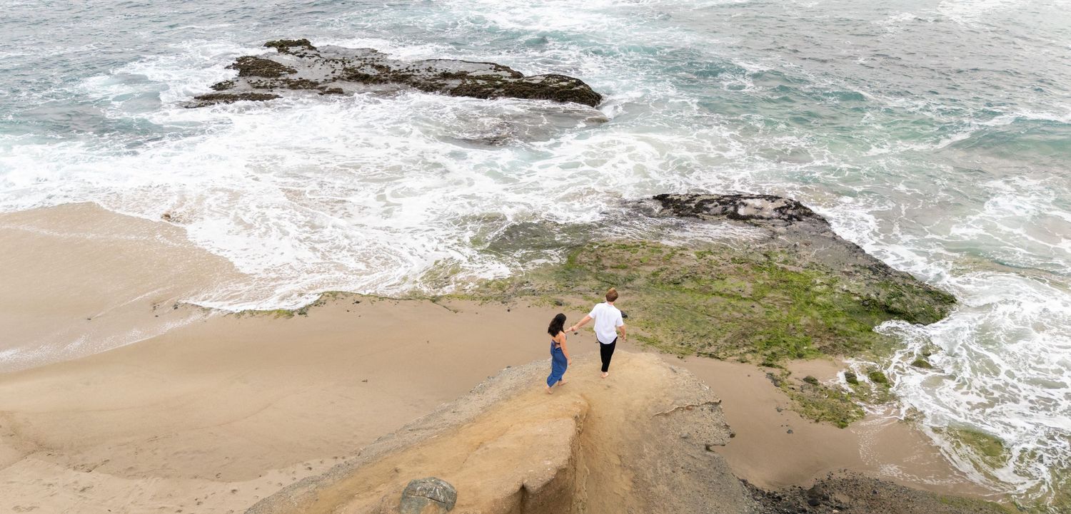 Two people walk along a scenic coastal cliff with crashing waves and sandy beach below.