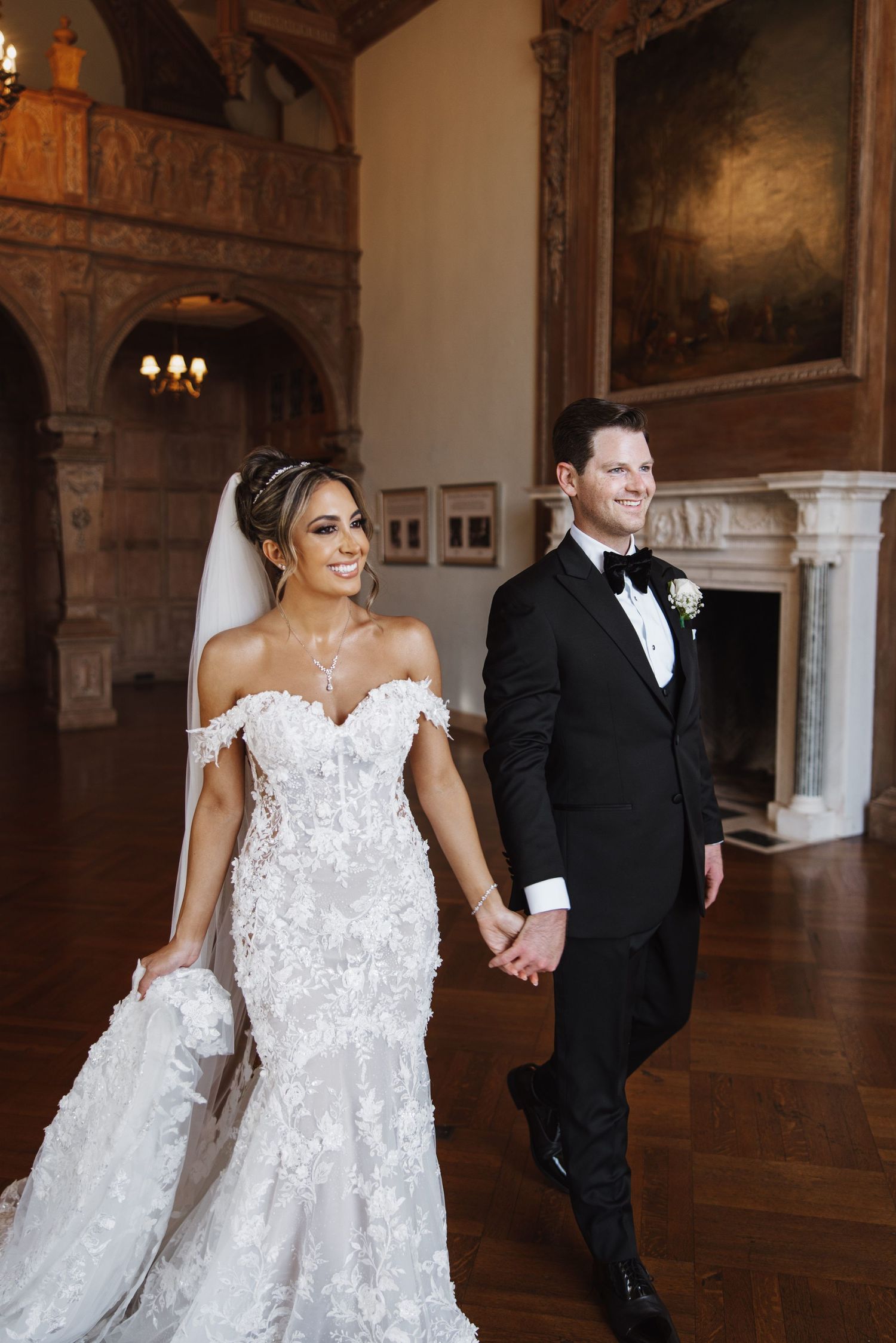 Couple in formal wedding attire walking through an elegant venue with columned corridor and chandeliers.