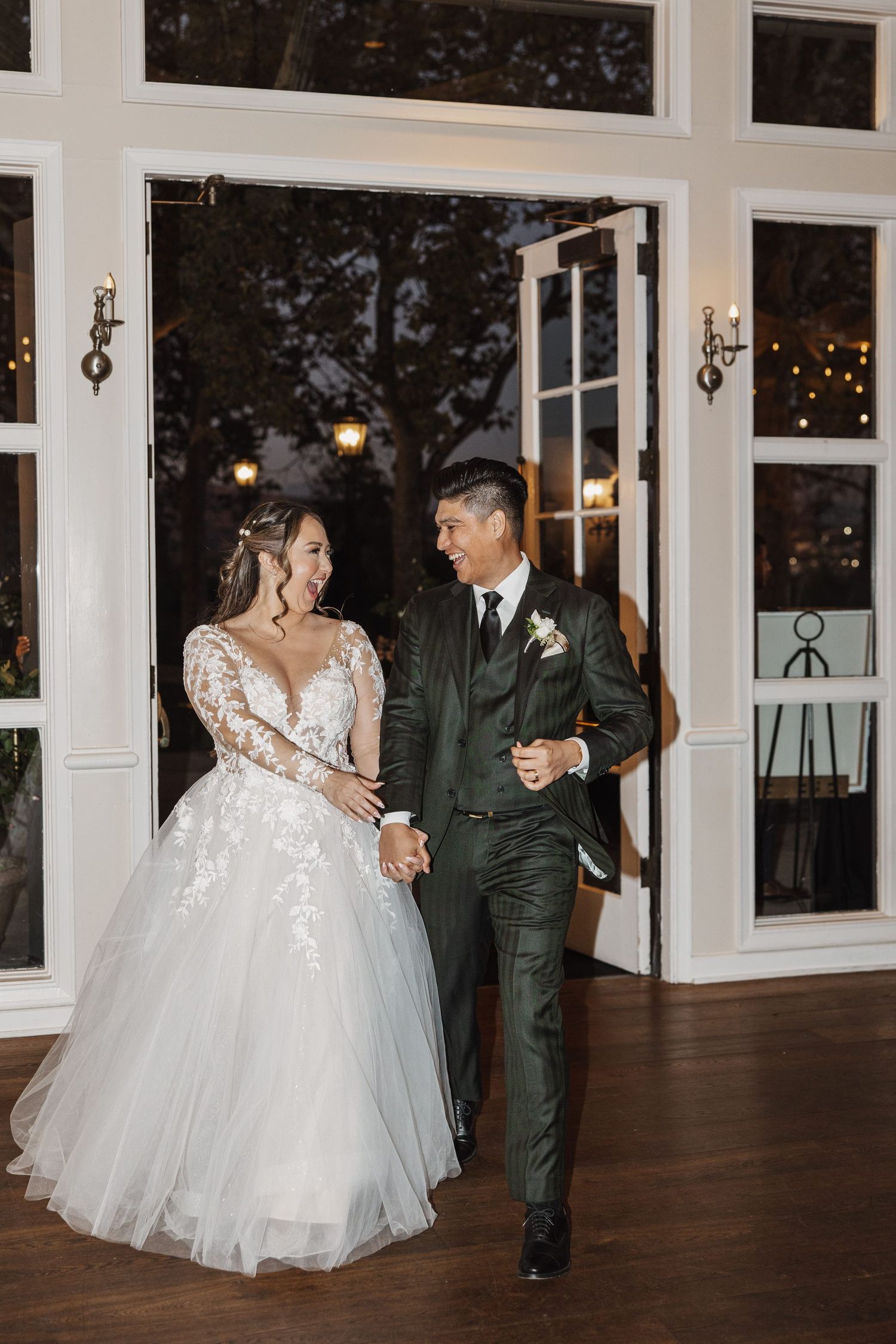 A couple in wedding attire share a joyful moment dancing together on a hardwood floor in an elegant venue with large windows.