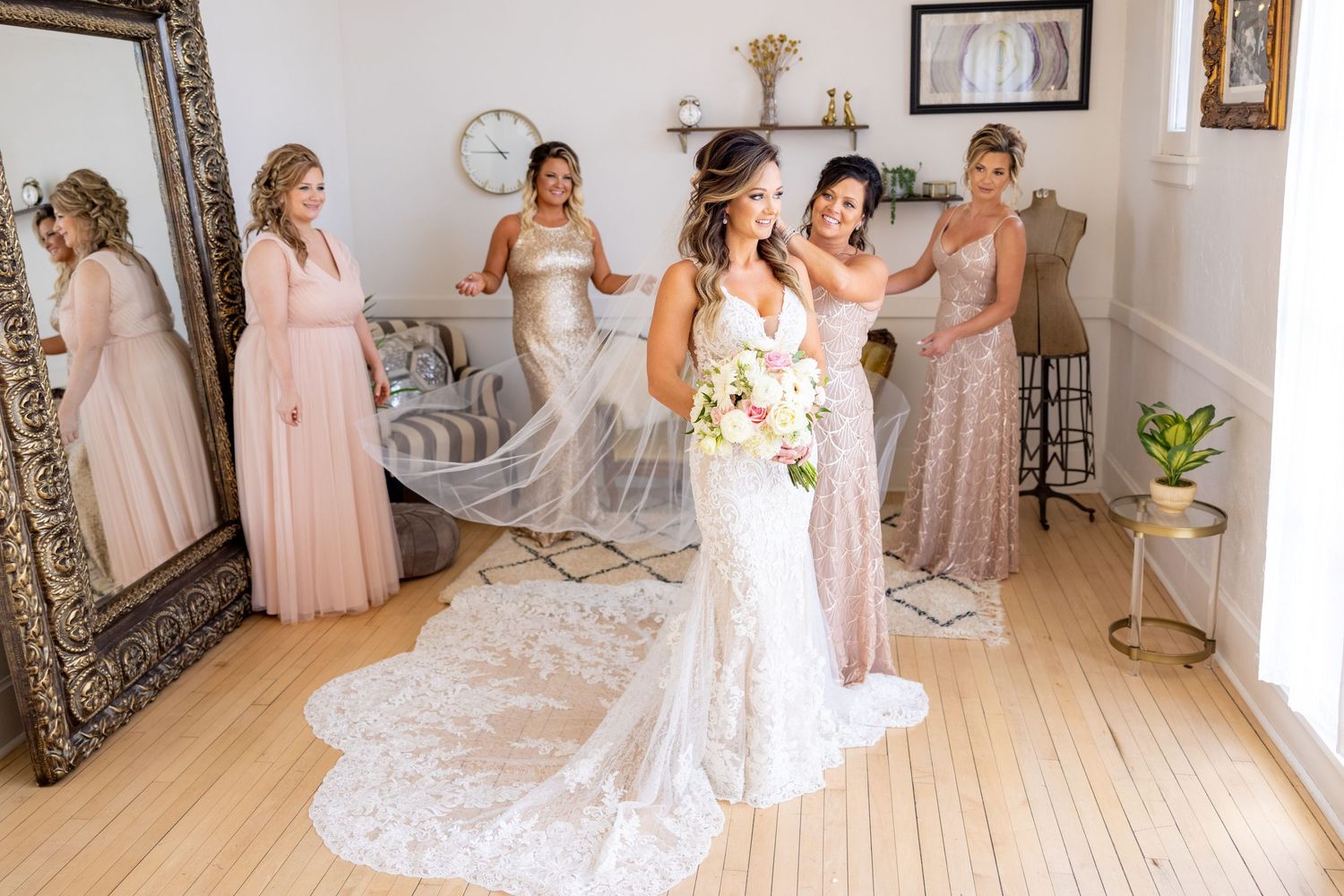 Bridesmaids in blush pink dresses help bride in lace wedding gown prepare in bright sunlit room.