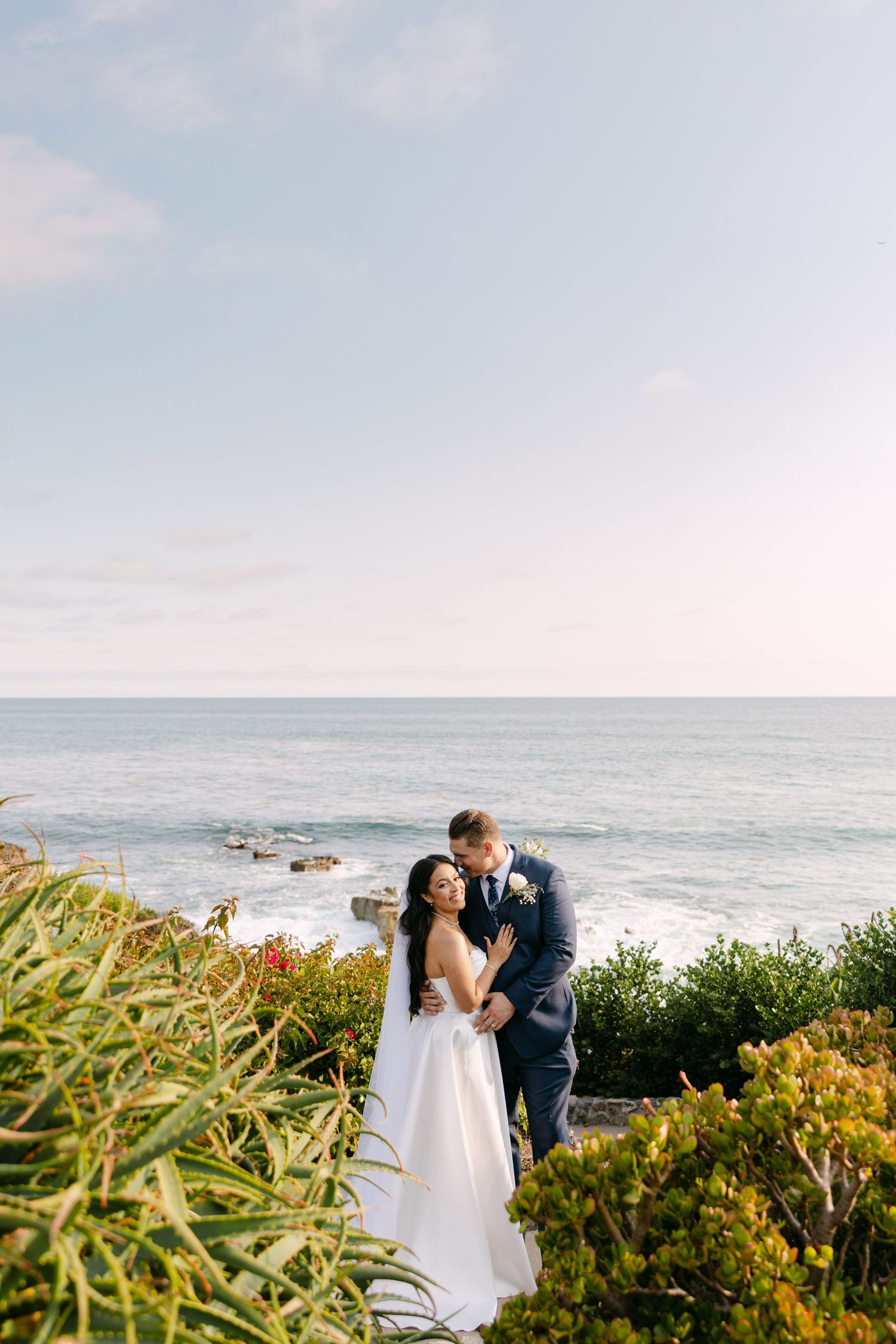 Newlywed couple shares romantic moments along a coastal cliff with ocean views and windswept grass in the foreground.