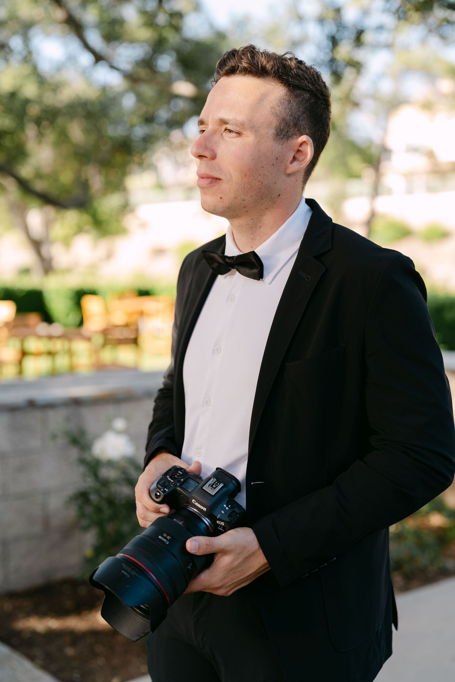Professional photographer in formal black tuxedo and bowtie holding camera with telephoto lens at outdoor event.