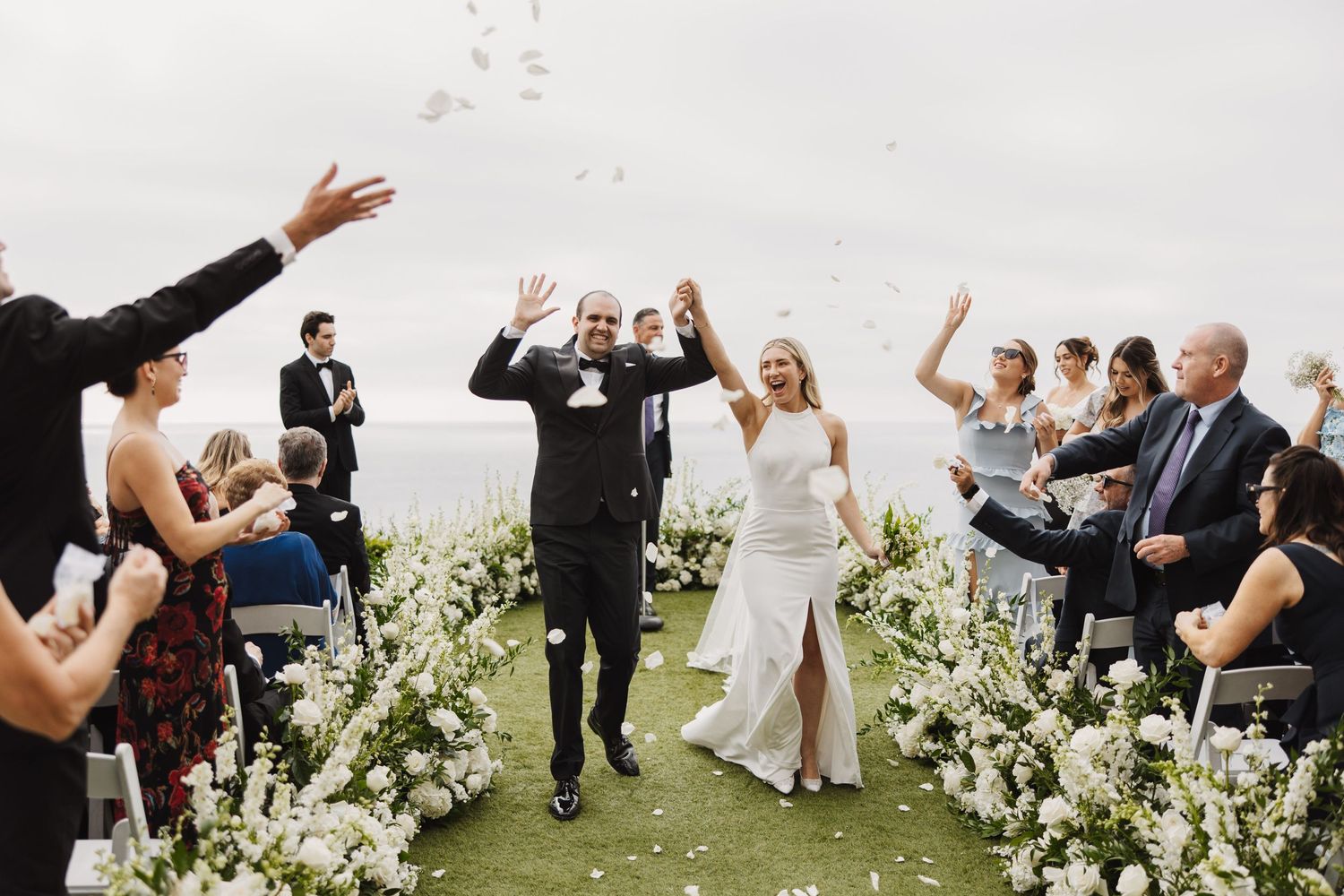 Wedding celebration with guests tossing flower petals during an outdoor ceremony surrounded by white floral arrangements.