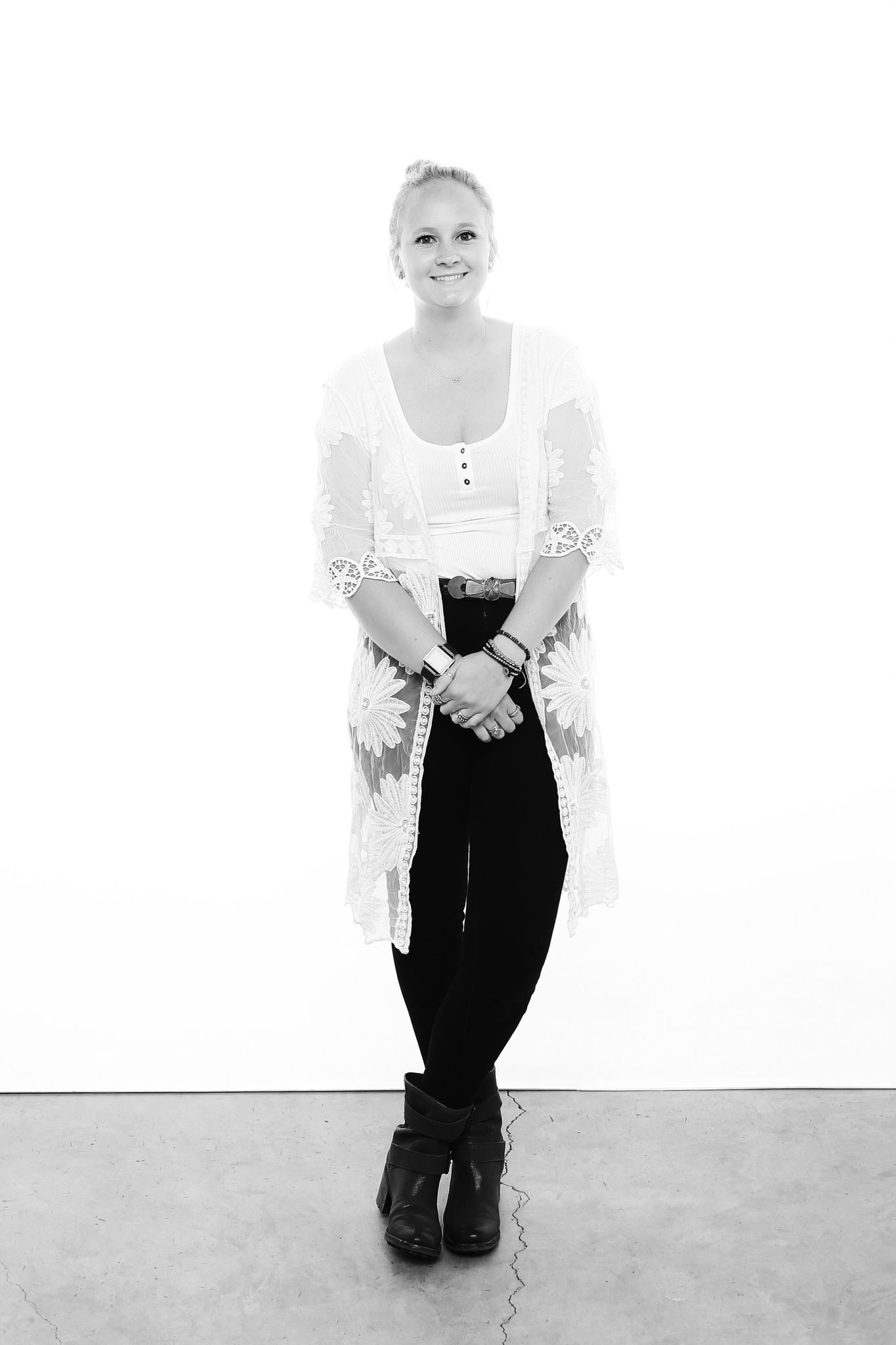 Black and white portrait of someone wearing a white lace cardigan over a tank top with black pants and boots against white wall.