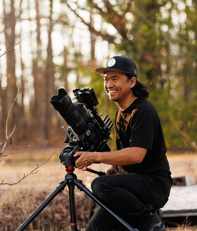 Photographer with camera equipment smiling while working outdoors in natural setting.