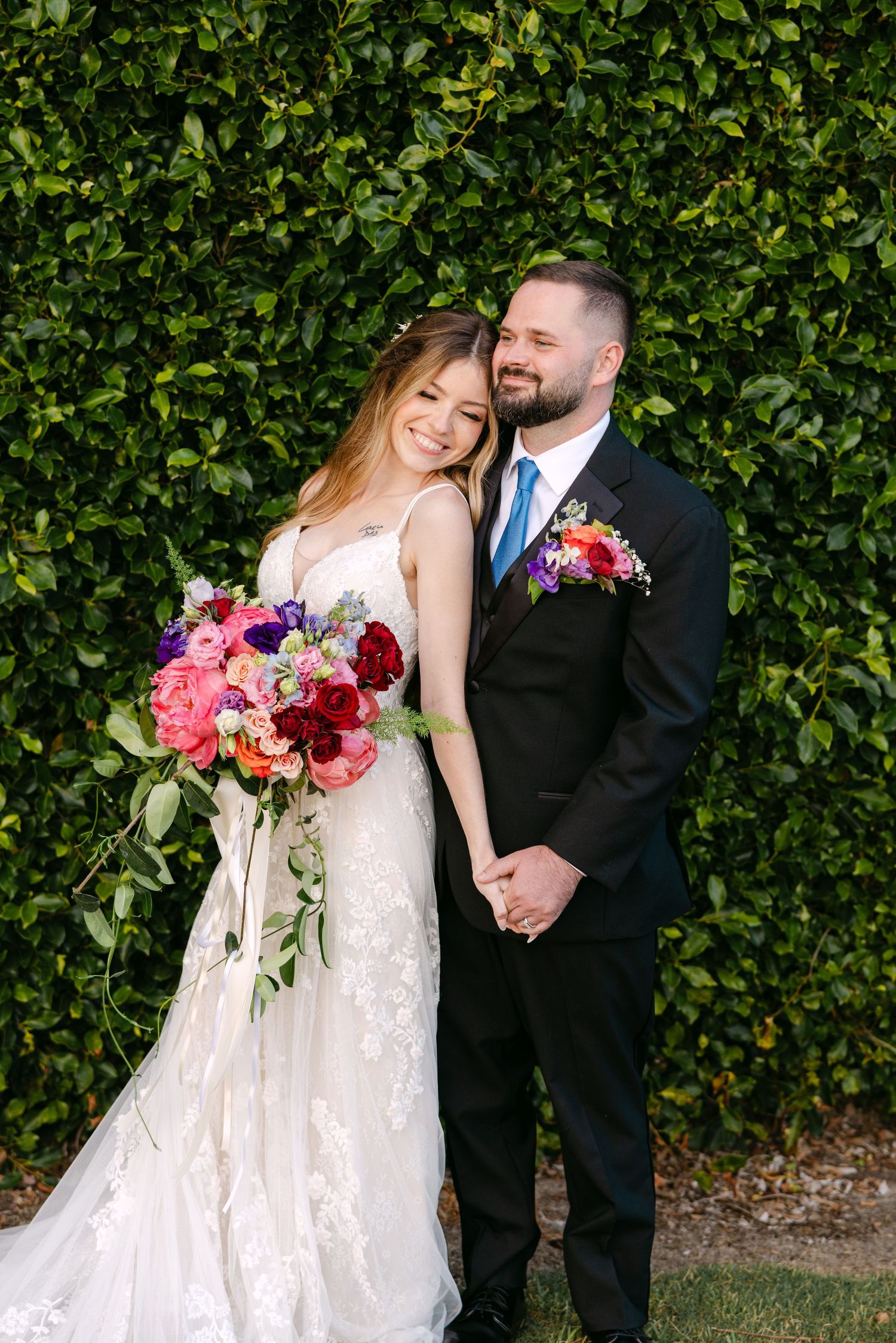 A couple in wedding attire poses in front of a lush green hedge wall, with a vibrant pink and red bouquet.