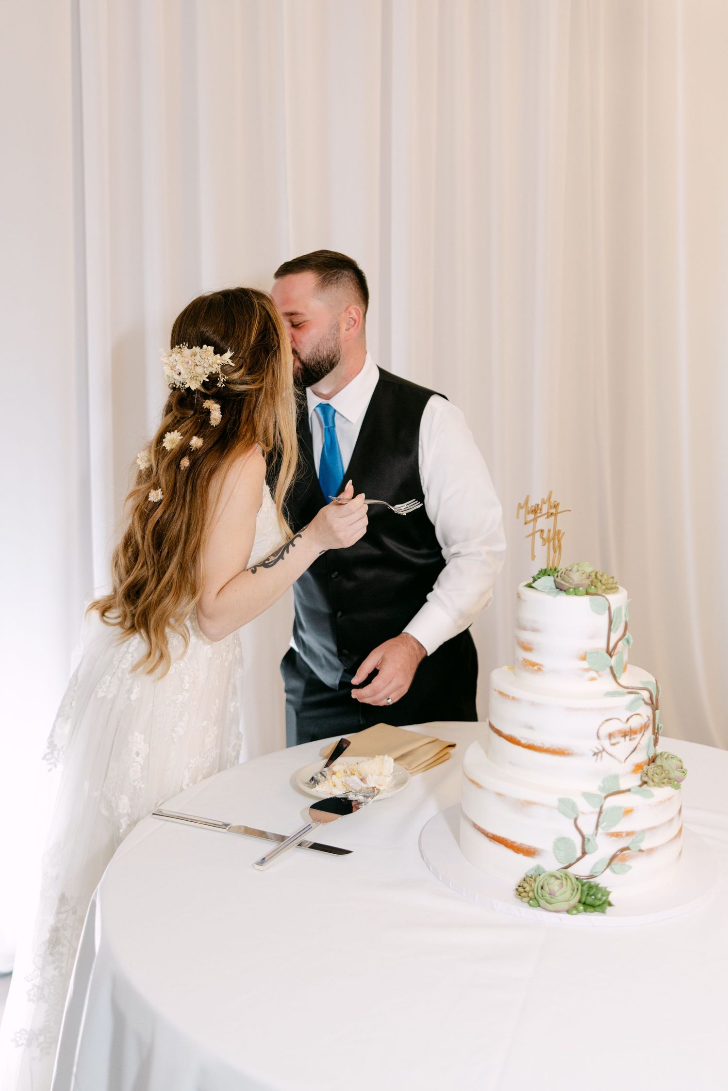 Newlyweds share an intimate moment while cutting their elegant three-tier white wedding cake adorned with greenery.