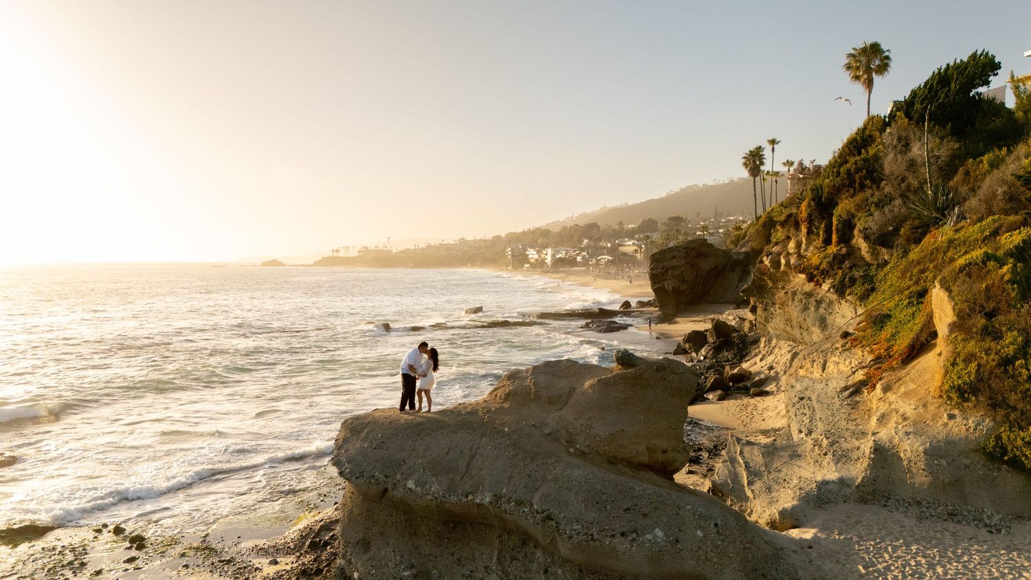 A surfer walks along a rocky beach at sunset in Laguna Beach, California with cliffs and ocean waves in view.