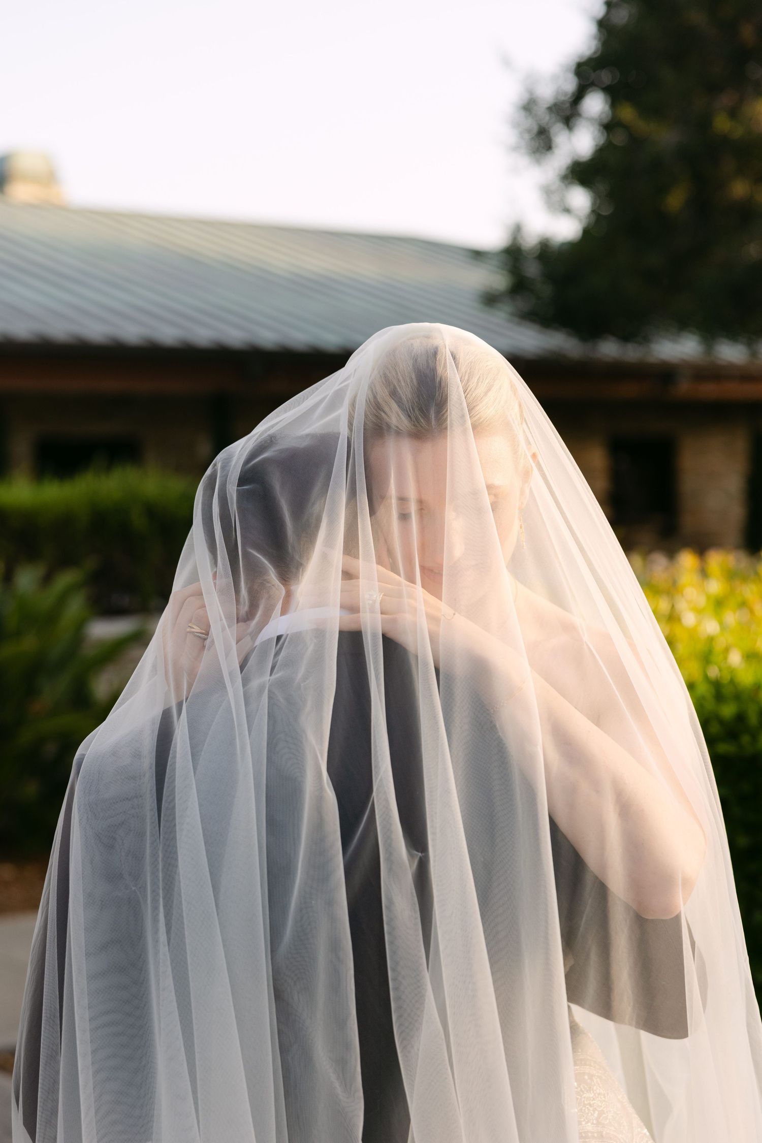 A bride wearing a delicate sheer veil that drapes over her figure stands outdoors in soft golden light.