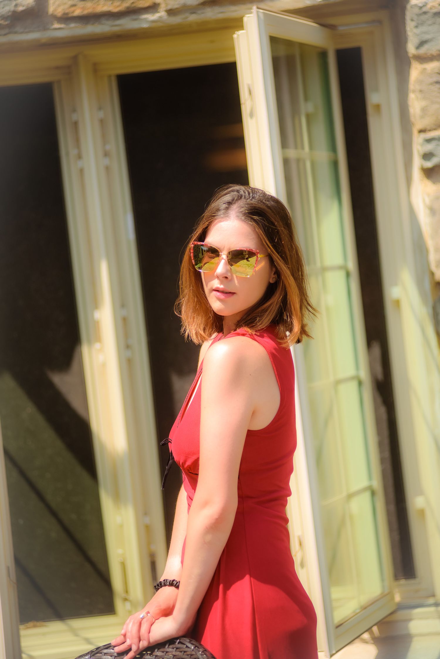 Young woman in red dress and yellow mirrored sunglasses posing outdoors near open French doors in sunlight.
