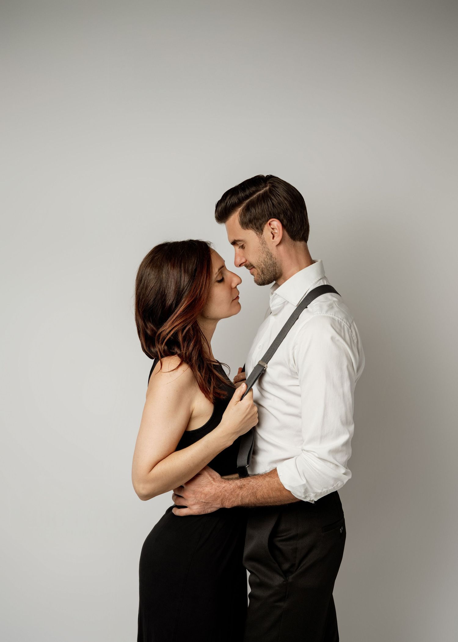 Romantic couple in elegant attire embrace intimately against neutral backdrop during stylish editorial photoshoot.