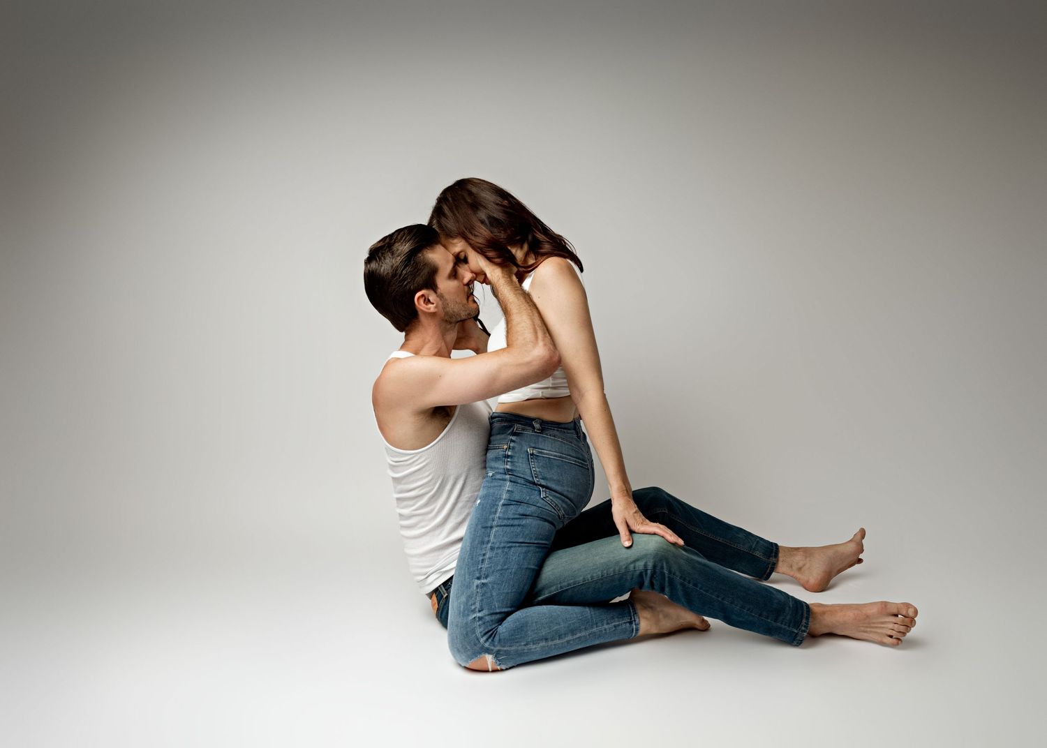 An intimate couple in casual attire embracing while sitting on a white floor against a neutral background.