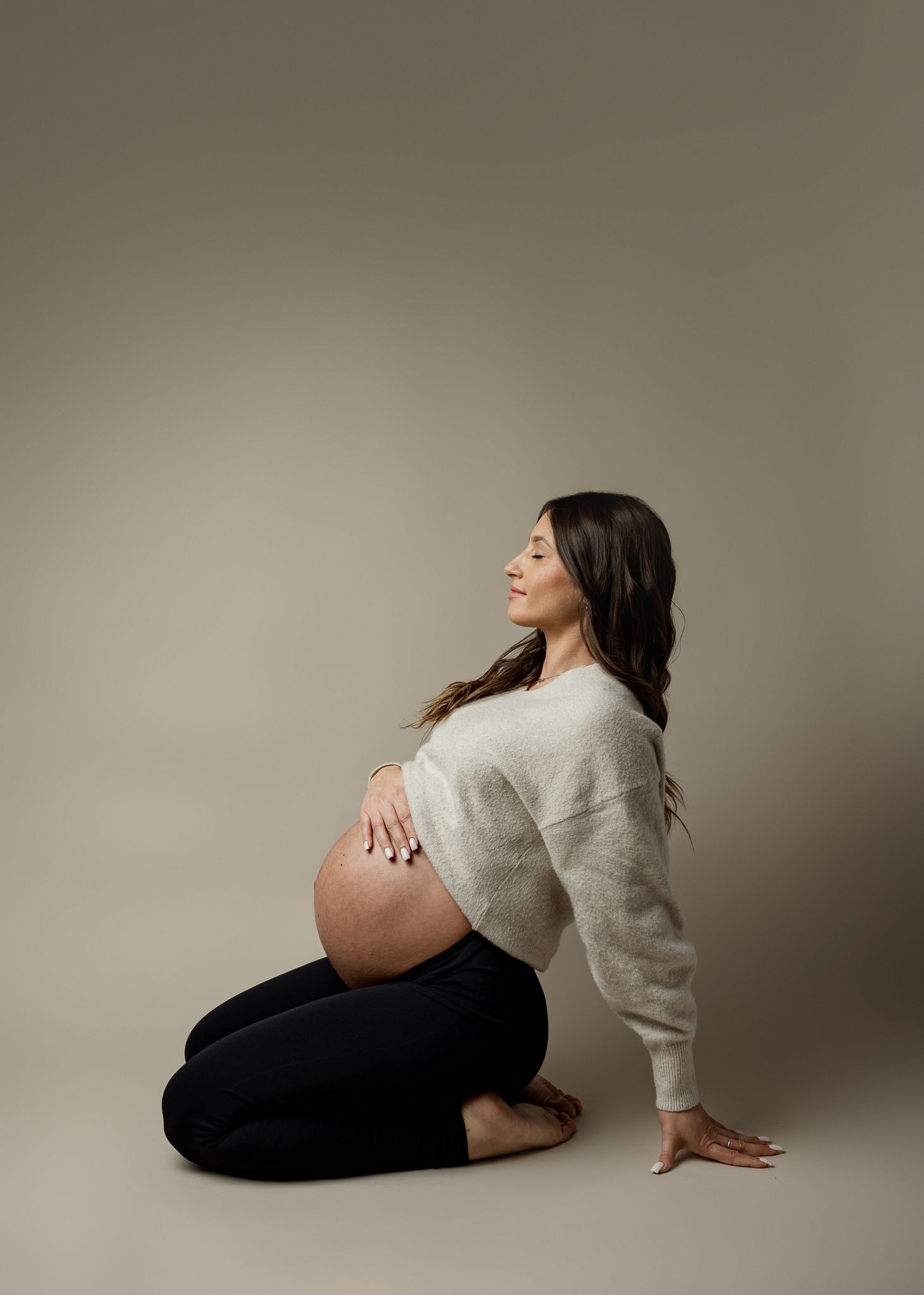 A pregnant woman in black pants and gray sweater poses gracefully on a neutral backdrop in an artistic maternity photoshoot.