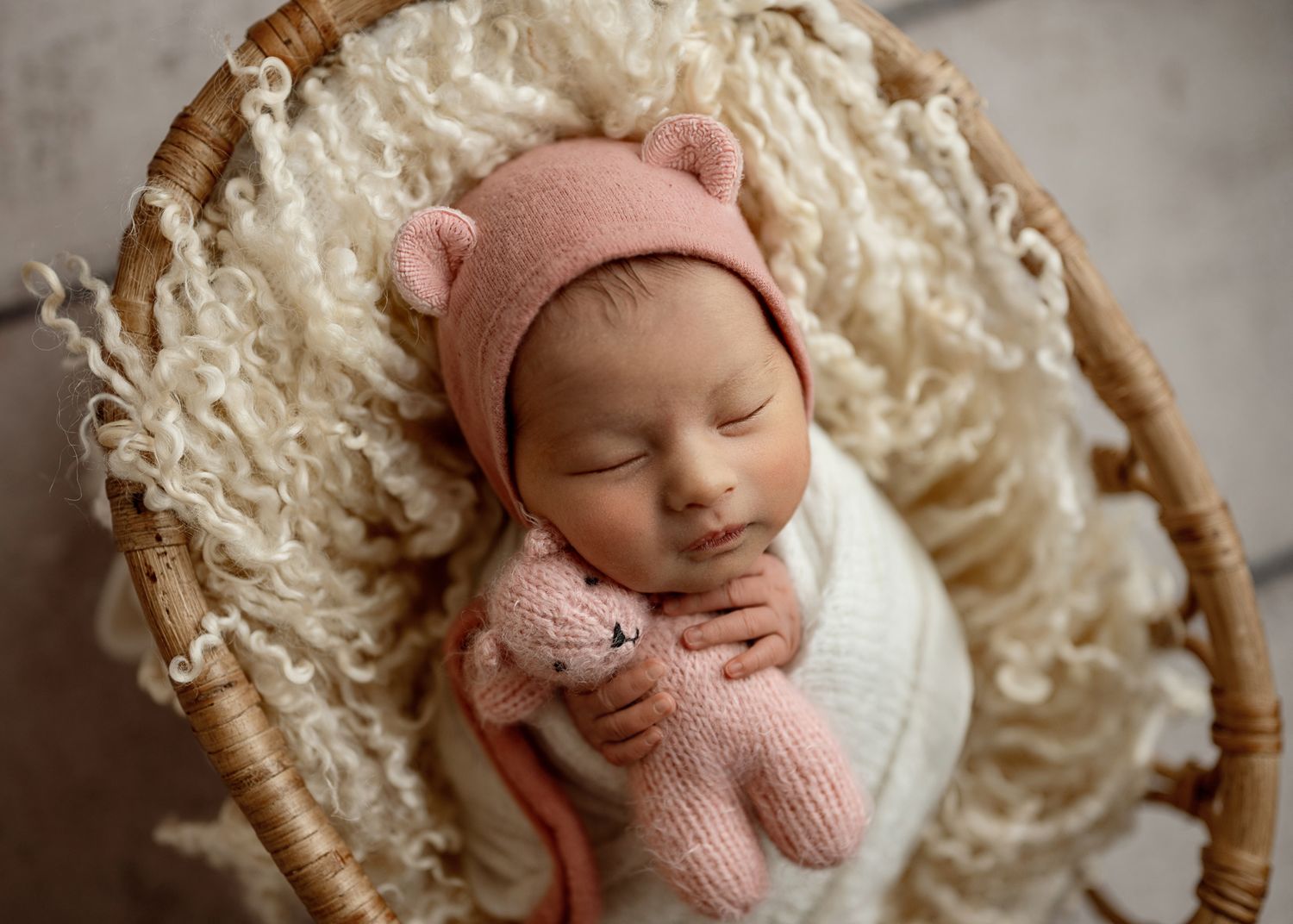 Newborn baby in pink hat sleeping peacefully in white textured basket.