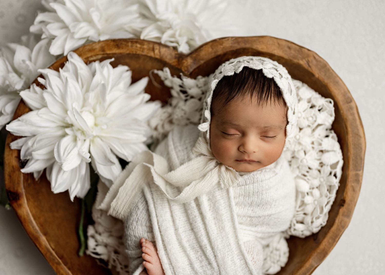 Sleeping newborn in white swaddle blanket nestled in heart-shaped wooden bowl with white flowers.