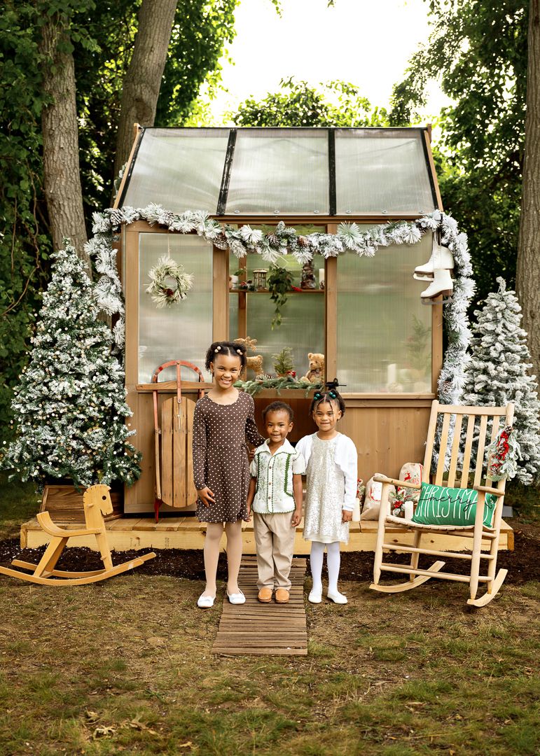 Three children pose in front of a decorated holiday greenhouse with flocked trees and garland in a backyard setting.