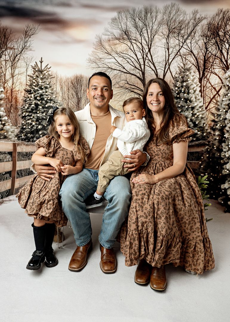 Family of four posing for Christmas portrait with snowy trees and wooden fence backdrop.