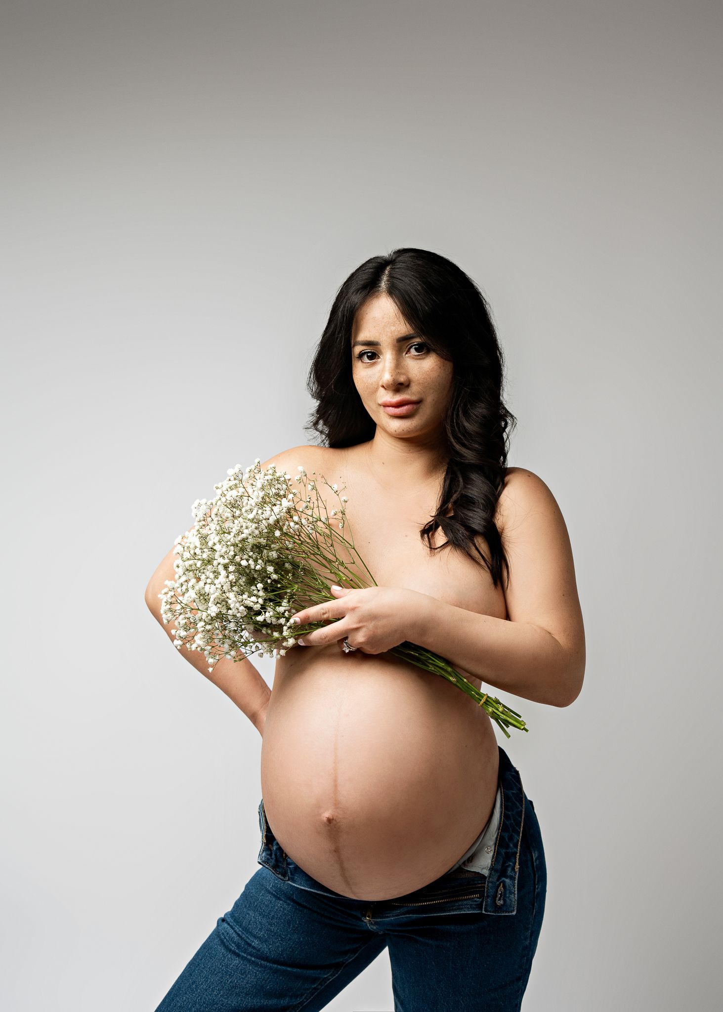 Pregnant woman in jeans holds a delicate bouquet of baby's breath flowers against a neutral background.