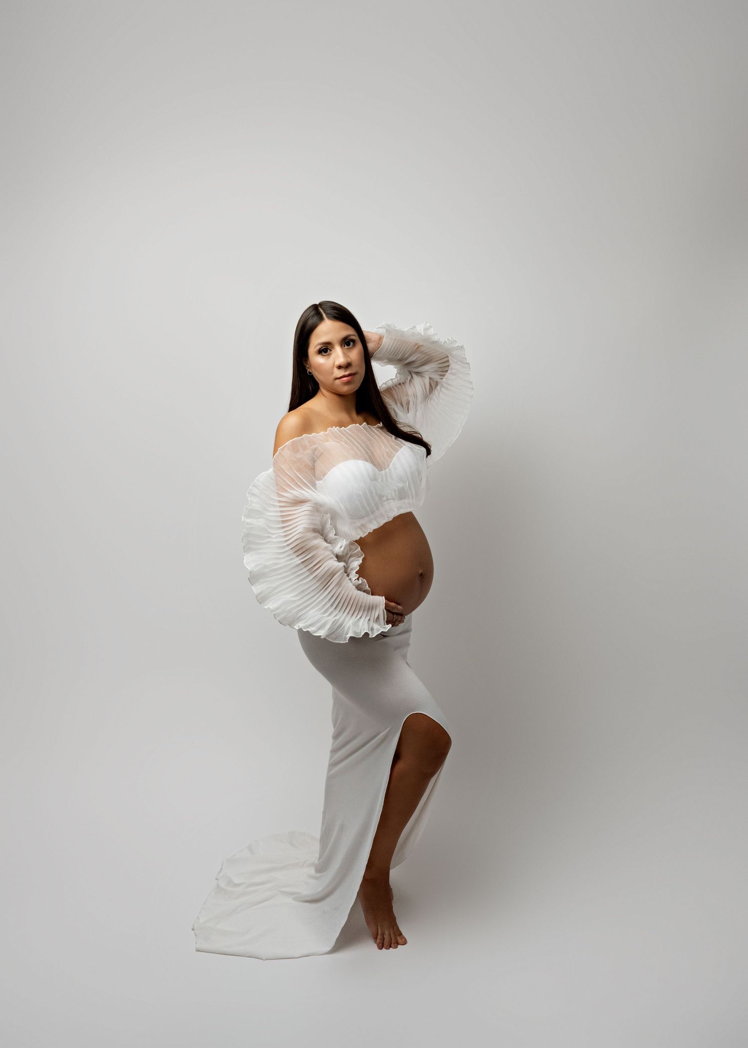 A figure in a flowing white dress poses gracefully against a light gray backdrop in an artistic portrait series.