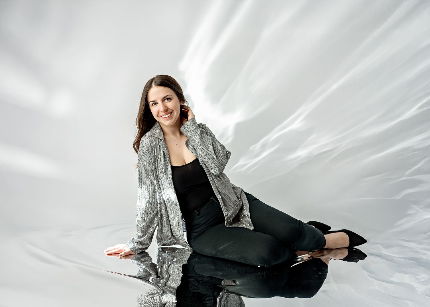 Professional studio portrait of a person in silver sequined jacket and black outfit sitting against white backdrop.
