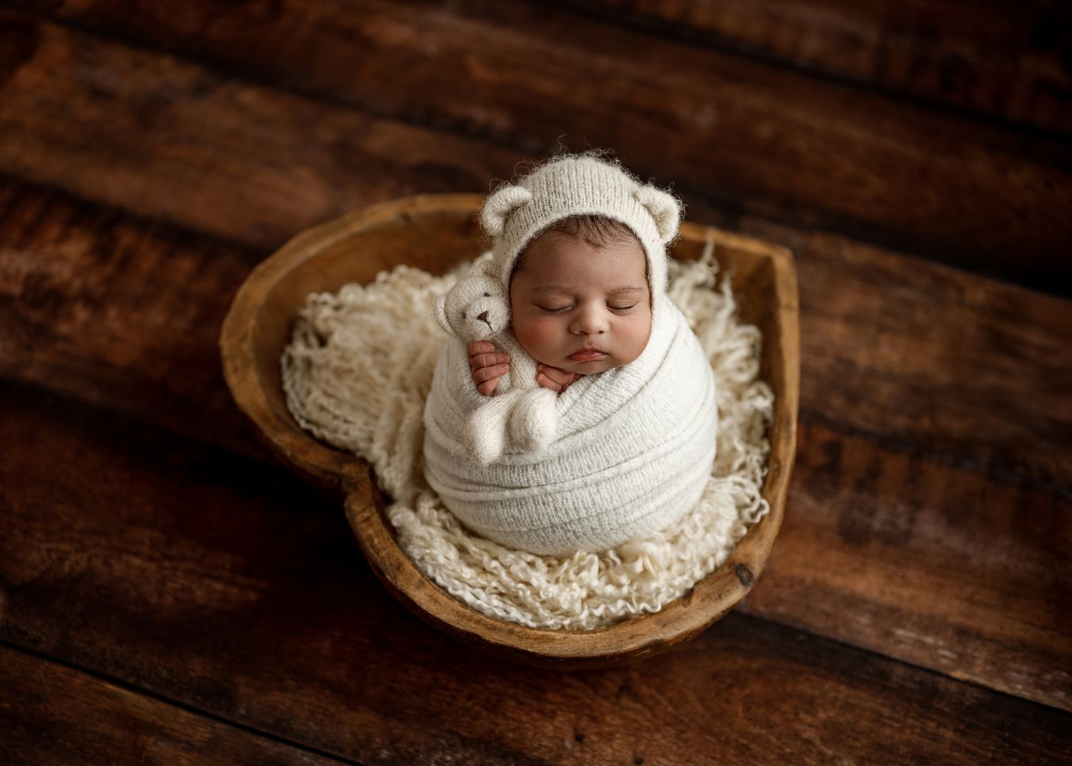 Sleeping baby wrapped in white knit blanket rests peacefully in wooden heart-shaped bowl on rustic wooden surface.